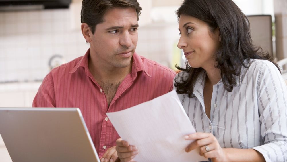 Couple in kitchen with paperwork using laptop looking unhappy