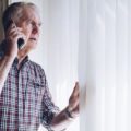Senior man talking on the phone whilst looking out of the window to his neighborhood. He has a worried look on his face do to security concerns.