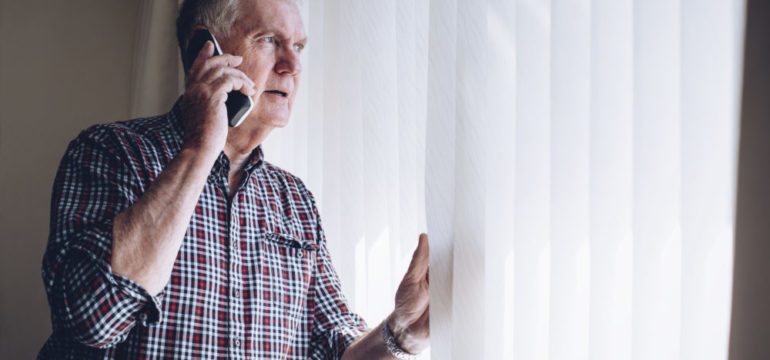 Senior man talking on the phone whilst looking out of the window to his neighborhood. He has a worried look on his face do to security concerns.