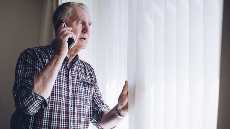 Senior man talking on the phone whilst looking out of the window to his neighborhood. He has a worried look on his face do to security concerns.