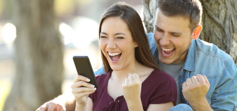 Excited couple reading good news in a smart phone that they won a bidding war sitting in a park