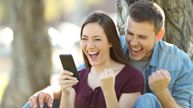 Excited couple reading good news in a smart phone that they won a bidding war sitting in a park
