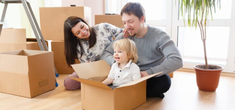 Happy young family with unpacking boxes at new home on moving day. Mother, father and little kid son having fun and smiling, relocation company concept.