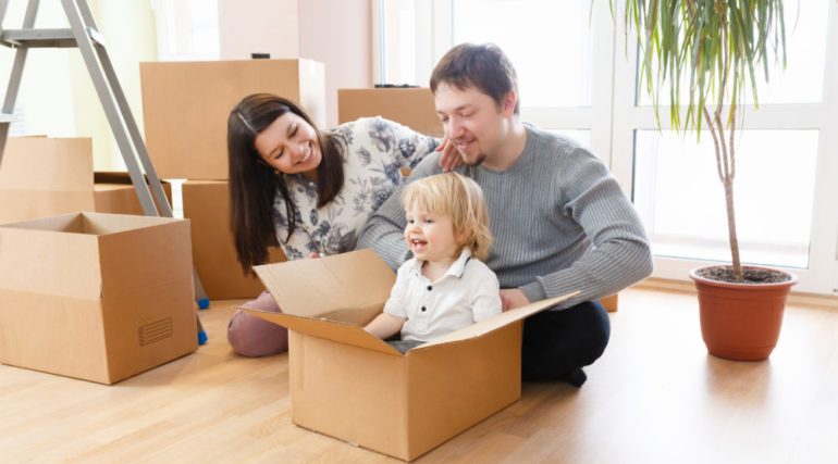 Happy young family with unpacking boxes at new home on moving day. Mother, father and little kid son having fun and smiling, relocation company concept.