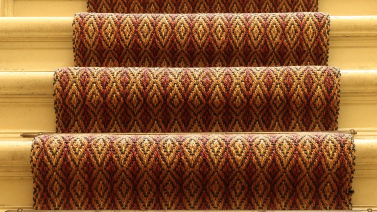 Textured carpet runners in red and gold pattern on light hardwood stairs.