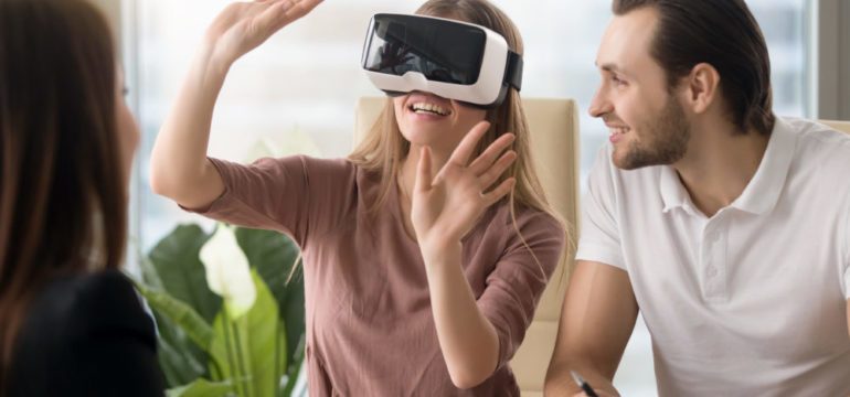 Business team of three people working on virtual reality applications and games, young excited woman testing VR glasses or goggles sitting in the office room with two colleagues, teleconference.
