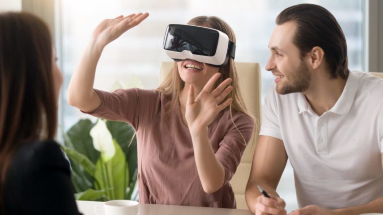 Business team of three people working on virtual reality applications and games, young excited woman testing VR glasses or goggles sitting in the office room with two colleagues, teleconference.