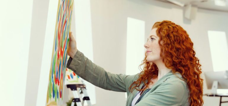 Young woman standing in an art gallery buying art. She is holding a colorful painting displayed against a white wall.