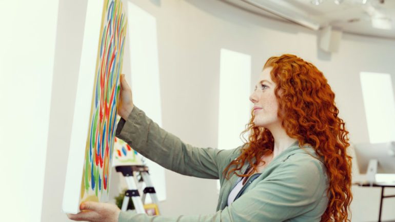 Young woman standing in an art gallery buying art. She is holding a colorful painting displayed against a white wall.