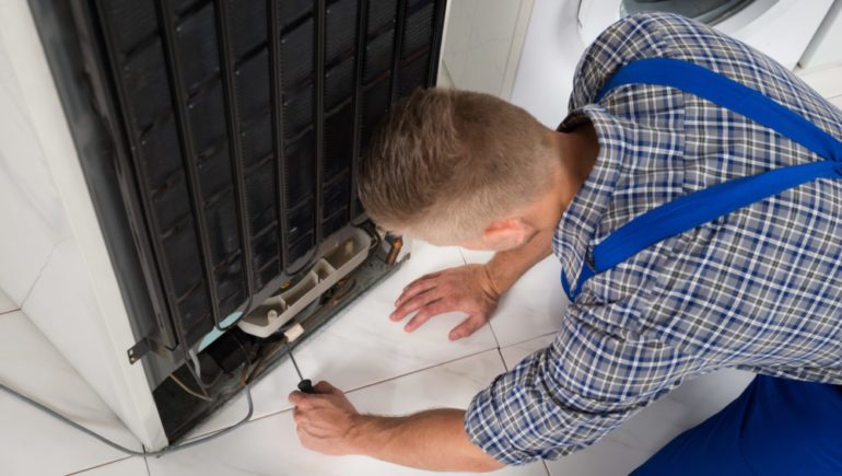 Young male repairman making repairs to home refrigeration appliance In kitchen.