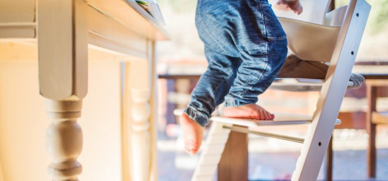Little toddler boy climbing on wooden highchair in danger of furniture toppling on him. Creates a dangerous situation for a young child in the home.