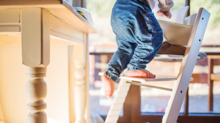 Little toddler boy climbing on wooden highchair in danger of furniture toppling on him. Creates a dangerous situation for a young child in the home.