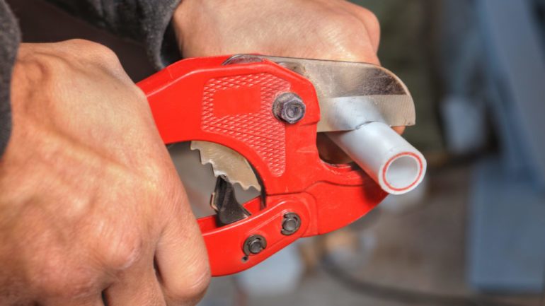 Polybutylene plumbing being cut by a plumber with a red pipe cutter.