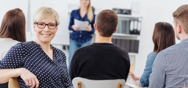 A happy female neighbor with glasses smiling at the camera during a homeowners association meeting with other members of the community.