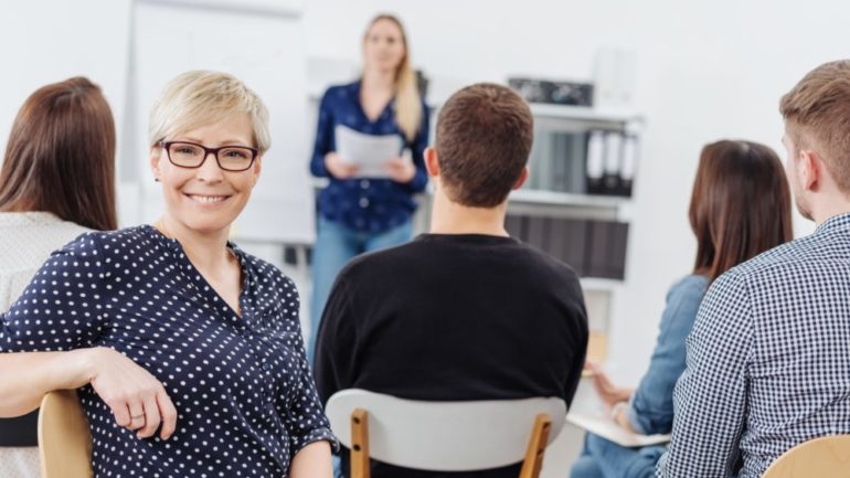 A happy female neighbor with glasses smiling at the camera during a homeowners association meeting with other members of the community.