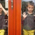 Two little boys wearing the same clothes looking through big glass storm and screen doors at the rain outside.