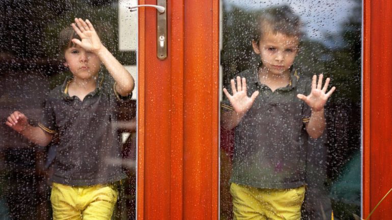 Two little boys wearing the same clothes looking through big glass storm and screen doors at the rain outside.