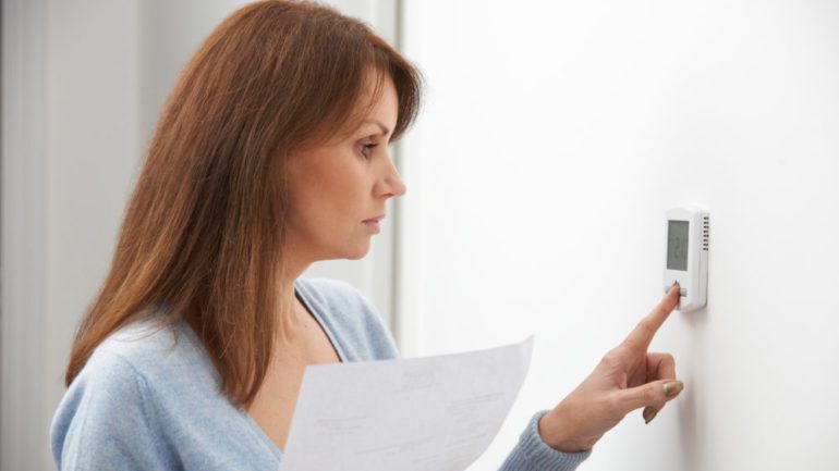 Worried woman turning down central heating thermostat with energy bill in hand.