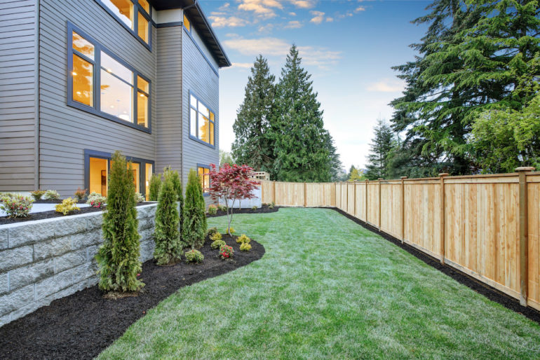 Luxurious contemporary three-story wood siding home exterior. Nice side yard landscape with well kept lawn, flower beds and wooden fence.