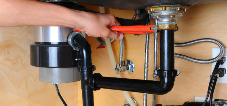 Closeup of a plumber using a wrench to tighten a fitting near a garbage disposal beneath a kitchen sink.