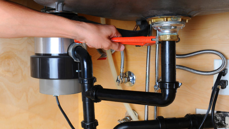 Closeup of a plumber using a wrench to tighten a fitting near a garbage disposal beneath a kitchen sink.