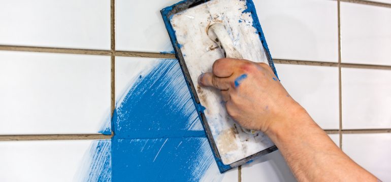 Builder applying colorful blue grout to newly laid white tiles on a wall using a rubber trowel to apply the mortar.