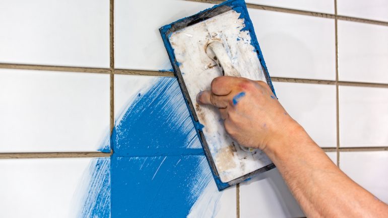 Builder applying colorful blue grout to newly laid white tiles on a wall using a rubber trowel to apply the mortar.