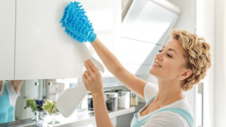 Busy house cleaning service worker is holding spray cleaner as she cleans a white kitchen cabinet.