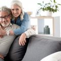Portrait of happy senior married couple relaxing together at home. Woman is standing and hugging man who is sitting on sofa. They are looking at camera and smiling thinking about where to retire.