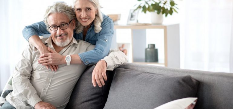 Portrait of happy senior married couple relaxing together at home. Woman is standing and hugging man who is sitting on sofa. They are looking at camera and smiling thinking about where to retire.