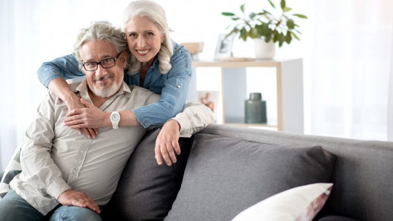 Portrait of happy senior married couple relaxing together at home. Woman is standing and hugging man who is sitting on sofa. They are looking at camera and smiling thinking about where to retire.