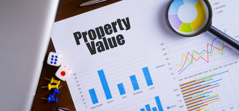 Property Value text on paper sheet may refer to a Zestimate with magnifying glass on chart, dice, spectacles, pen, laptop and blue and yellow push pin on wooden table.