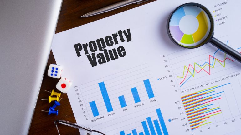 Property Value text on paper sheet may refer to a Zestimate with magnifying glass on chart, dice, spectacles, pen, laptop and blue and yellow push pin on wooden table.