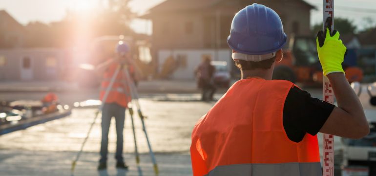 Property survey for new construction is being performed by two professionals wearing safety hard hats and vests.