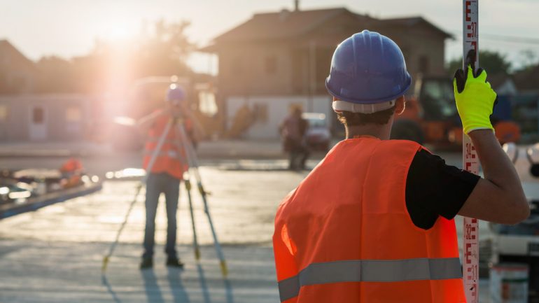 Property survey for new construction is being performed by two professionals wearing safety hard hats and vests.