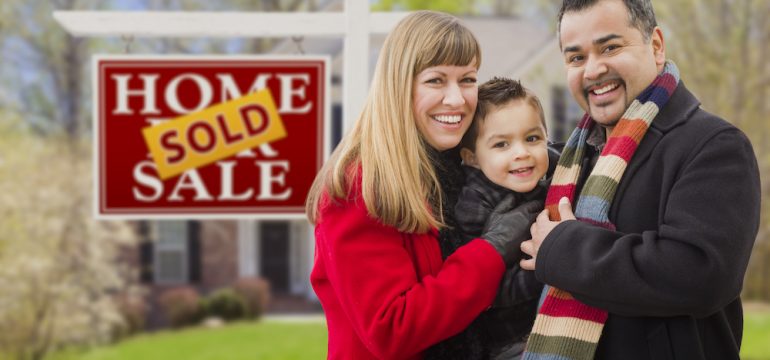 Warmly dressed young family in front of their home with Sold sticker over Home For Sale real estate sign. Represents selling your home in winter.