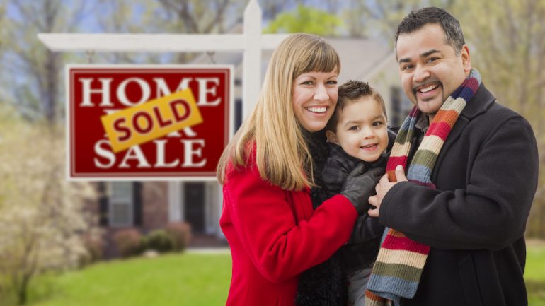 Warmly dressed young family in front of their home with Sold sticker over Home For Sale real estate sign. Represents selling your home in winter.