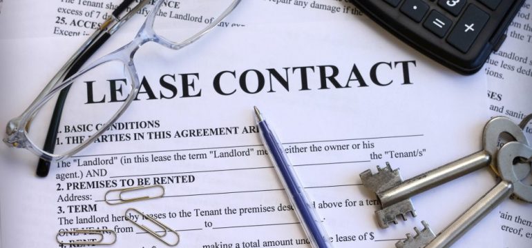 Close up copy of a lease contract that one would manage when buying a rental property with existing tenants. Eye glasses, a calculator, pen and house keys are also pictured on top of the contract.
