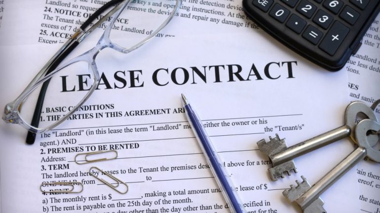 Close up copy of a lease contract that one would manage when buying a rental property with existing tenants. Eye glasses, a calculator, pen and house keys are also pictured on top of the contract.