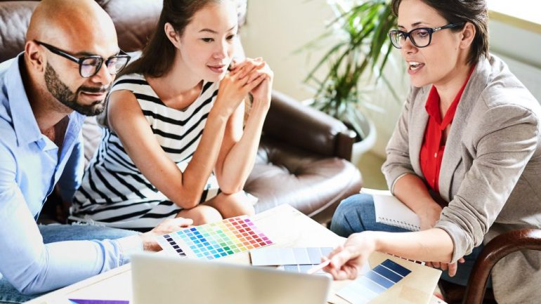 Pretty middle-aged interior decorator sharing ideas with her clients while having meeting in cozy boardroom, they listening to her with interest.