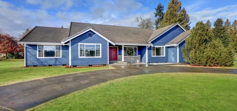 Beautiful rambler house with blue siding and covered porch with double red front door and concrete driveway on a large lot. Shows the buyer's decision to purchase a large lot size versus house size.
