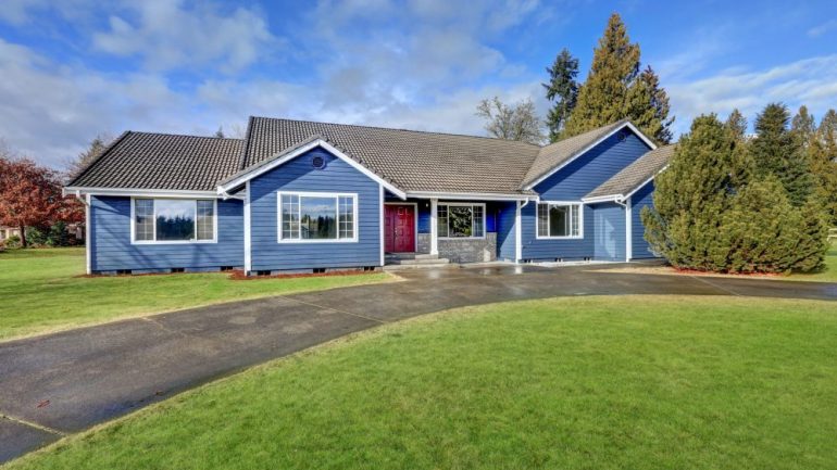 Beautiful rambler house with blue siding and covered porch with double red front door and concrete driveway on a large lot. Shows the buyer's decision to purchase a large lot size versus house size.