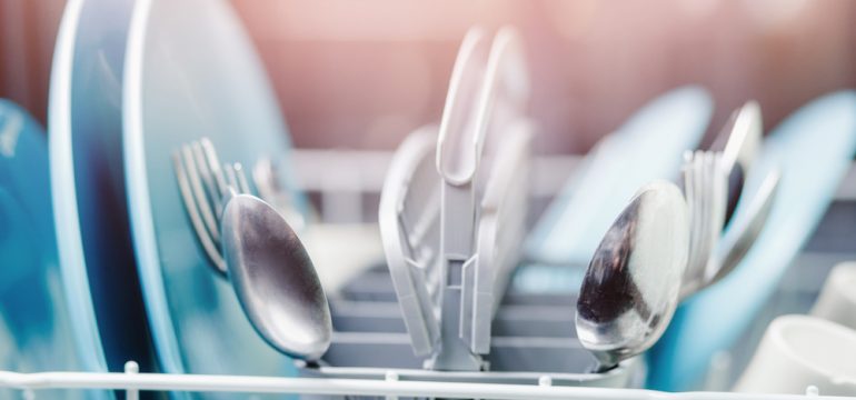 A dishwasher is a workhorse during the coronavirus pandemic. Spoons in dishwasher at foreground, background is blue, blurred dishes.