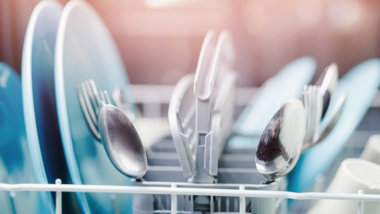 A dishwasher is a workhorse during the coronavirus pandemic. Spoons in dishwasher at foreground, background is blue, blurred dishes.