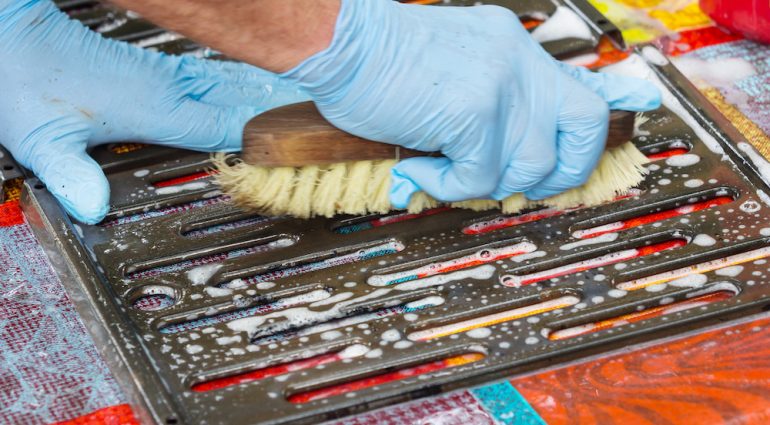 Grill cleaning with blue gloved hands scrubbing grates with a brush.