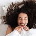 Portrait from above of cheerful woman with dark curly hair and a smiling face lying in bed under white bed sheets pulled up to her chin.