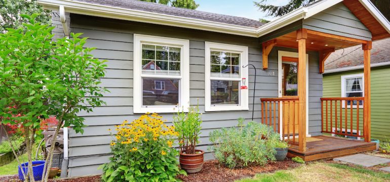 Small grey starter home with wooden deck. Front yard with flower bed and lawn.