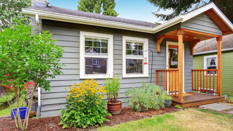 Small grey starter home with wooden deck. Front yard with flower bed and lawn.