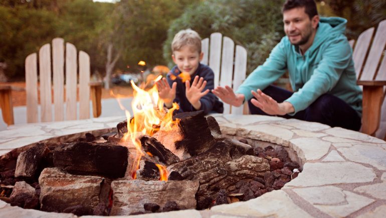View of firepit and happy smiling family of two, father and son, warming their hands by the fire and enjoying time together in the background.
