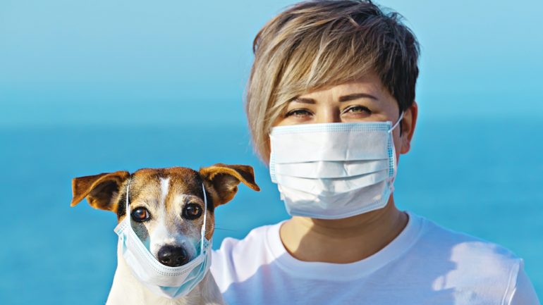 Woman in protective surgical mask holds dog in face mask. Coronavirus and pet care: safe practices during the COVID-19 pandemic.